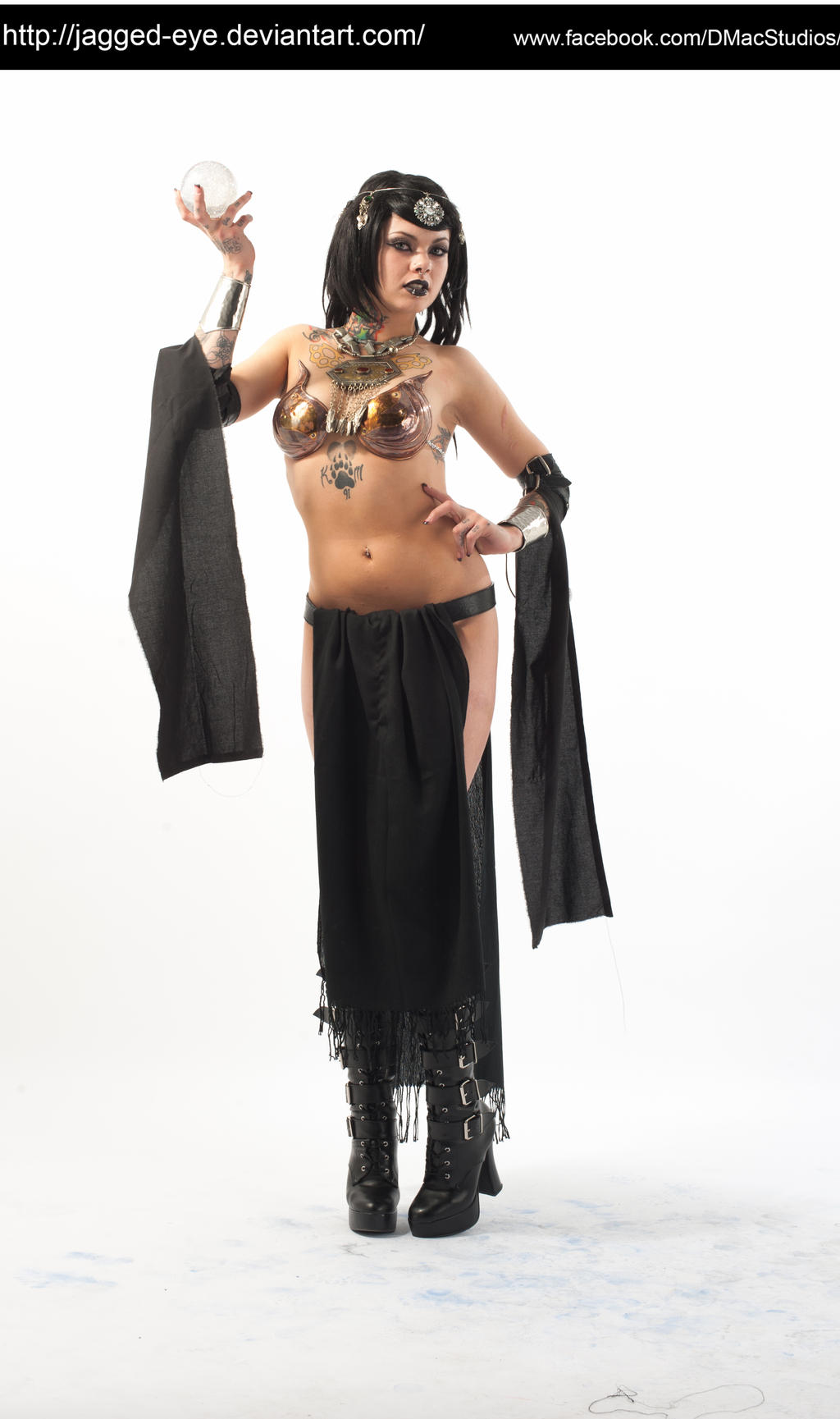 a full shot of a female model dressed in a dark fantasy costume.  she's wearing a black robelike garment over a bronzecolored bra and dark skirt.  she has dark hair and makeup, jewelry, and tattoos, and is holding a crystal ball. she's posed in a studio setting against a white backdrop. the overall style is dark fantasy or gothic, suggestive of a sorceress or belly dancer.