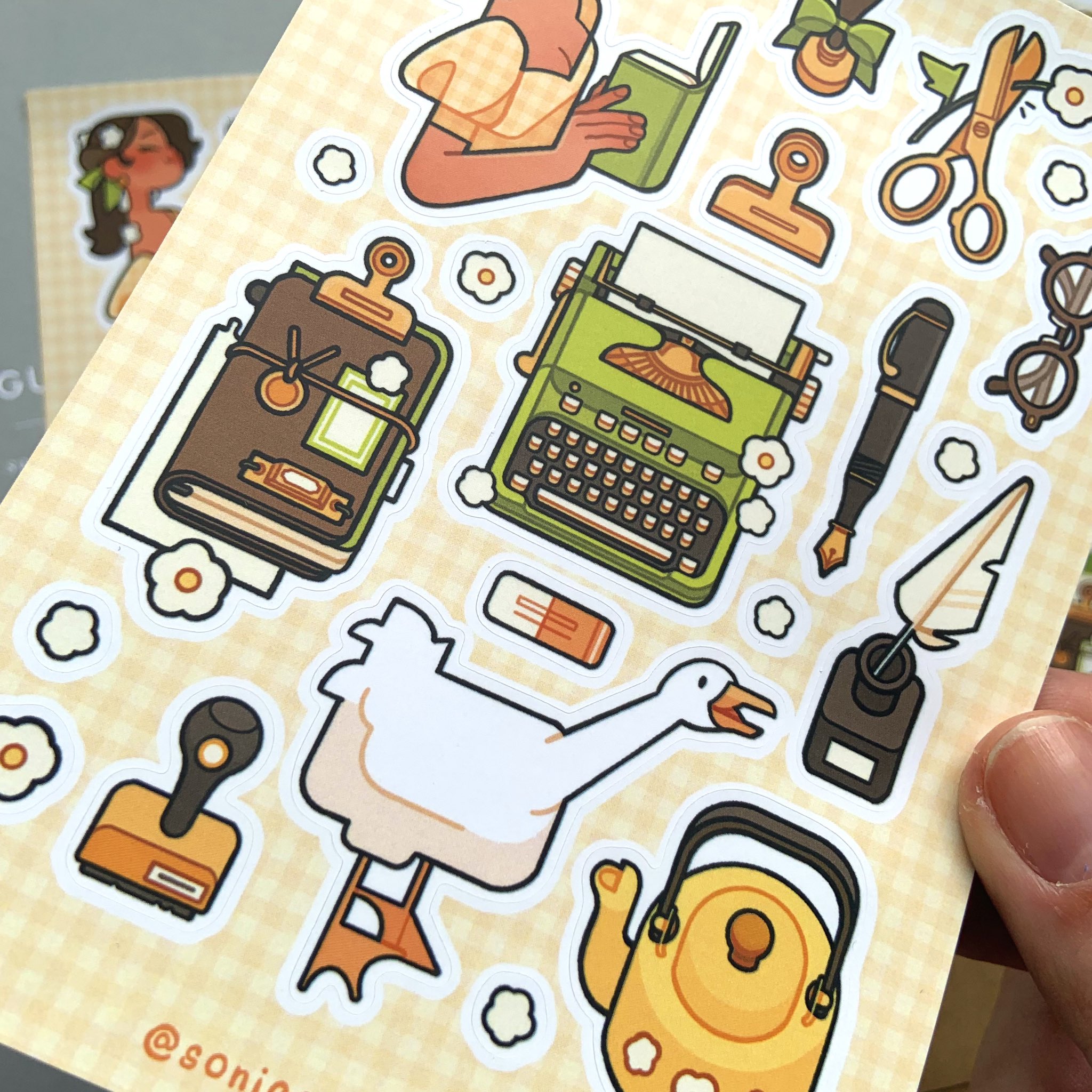 a hand holds a sheet of stickers with a vintage and cottagecore theme. the stickers feature illustrations of vintage styled stationary, plants, a woman, and a goose. the stickers are arranged on a pale yellow gingham background.