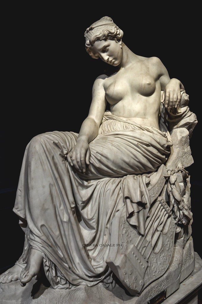 a classical marble statue of a nude female figure, possibly a greek or roman goddess, is illuminated against a black background. the statue captures the woman in a seated pose, her head slightly bowed, with drapery flowing over her lower body.  her left hand rests gracefully on her lap, while her right hand reaches back to rest on a broken column. a lyre, a musical instrument associated with poetry and music, lies at her feet. the studio lighting accentuates the smooth texture of the marble and the carefully sculpted details of the figure's anatomy.  the black background isolates the statue and draws the viewer's attention to its form and beauty.