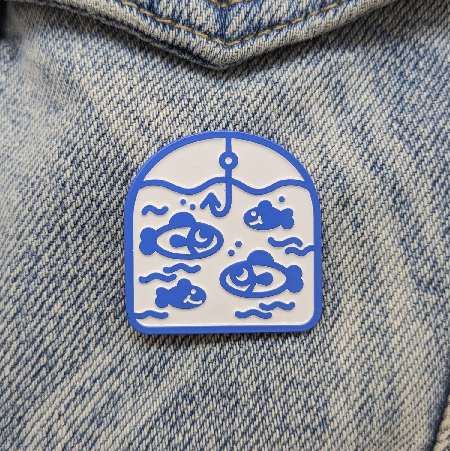 a closeup shot shows a blue and white enamel pin depicting a fishing scene. the pin features a blue outline and a white background. inside the pin are four stylized blue fish swimming among wavy lines, representing water. a fishing hook hangs from the top center of the pin. the pin is attached to a piece of denim fabric, likely a jacket or bag.