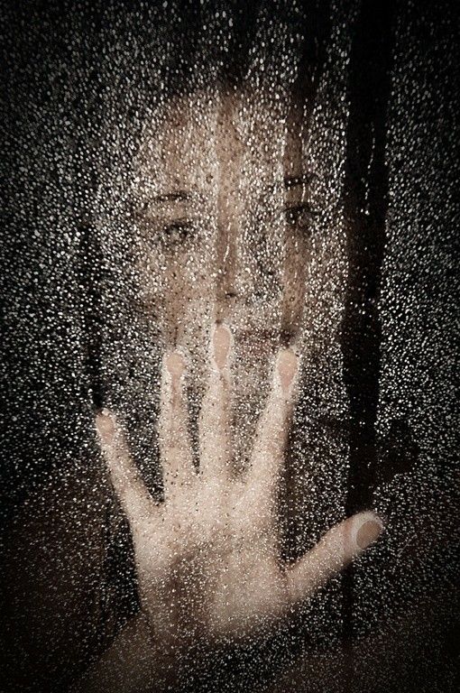 the image is a closeup, darkly lit portrait of a woman's face and hand pressed against a wet, possibly glass surface.  water droplets heavily obscure the view, creating a blurry and somewhat mysterious effect. the woman's expression appears to be one of sadness or despair, further emphasized by the dark and moody atmosphere.  her hand partially obscures her face, adding to the sense of concealment or introspection. the overall tone is somber and evokes feelings of isolation or loneliness. the use of water droplets adds a layer of texture and visual interest.  the colors are muted, giving a sepia or monochrome feel.