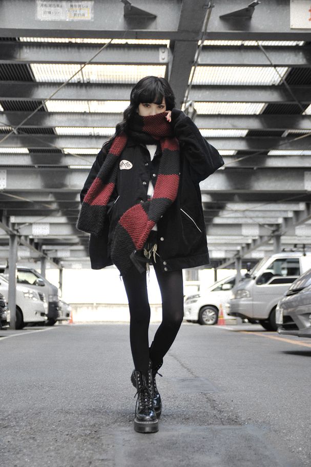 a young asian woman is stylishly dressed in dark clothing, posing in a parking garage. her outfit consists of a black overshirt or jacket, black tights, and black platform boots. she's wearing a striking red and black scarf that partially covers her face.  the overall aesthetic is grunge or gothinspired, with a dark and moody atmosphere enhanced by the setting of a grey parking garage.  the image emphasizes the model's fashion choices and overall style.