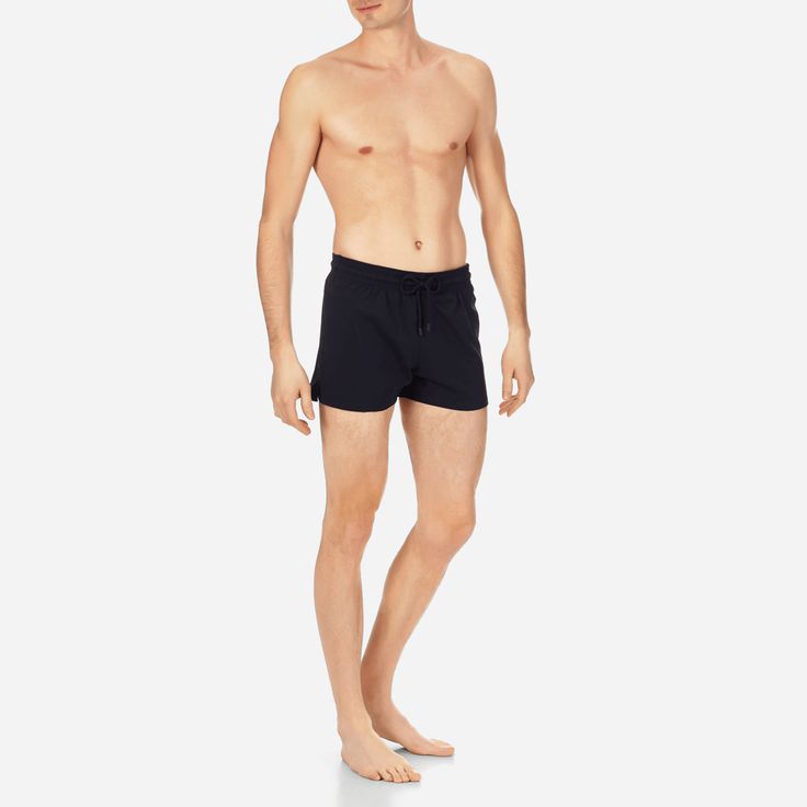 a man wearing black swim trunks is posing in a studio setting with a white background. he is standing with his legs slightly apart and his arms at his sides. the man has a toned physique.  he appears to be in his 20s or 30s.
