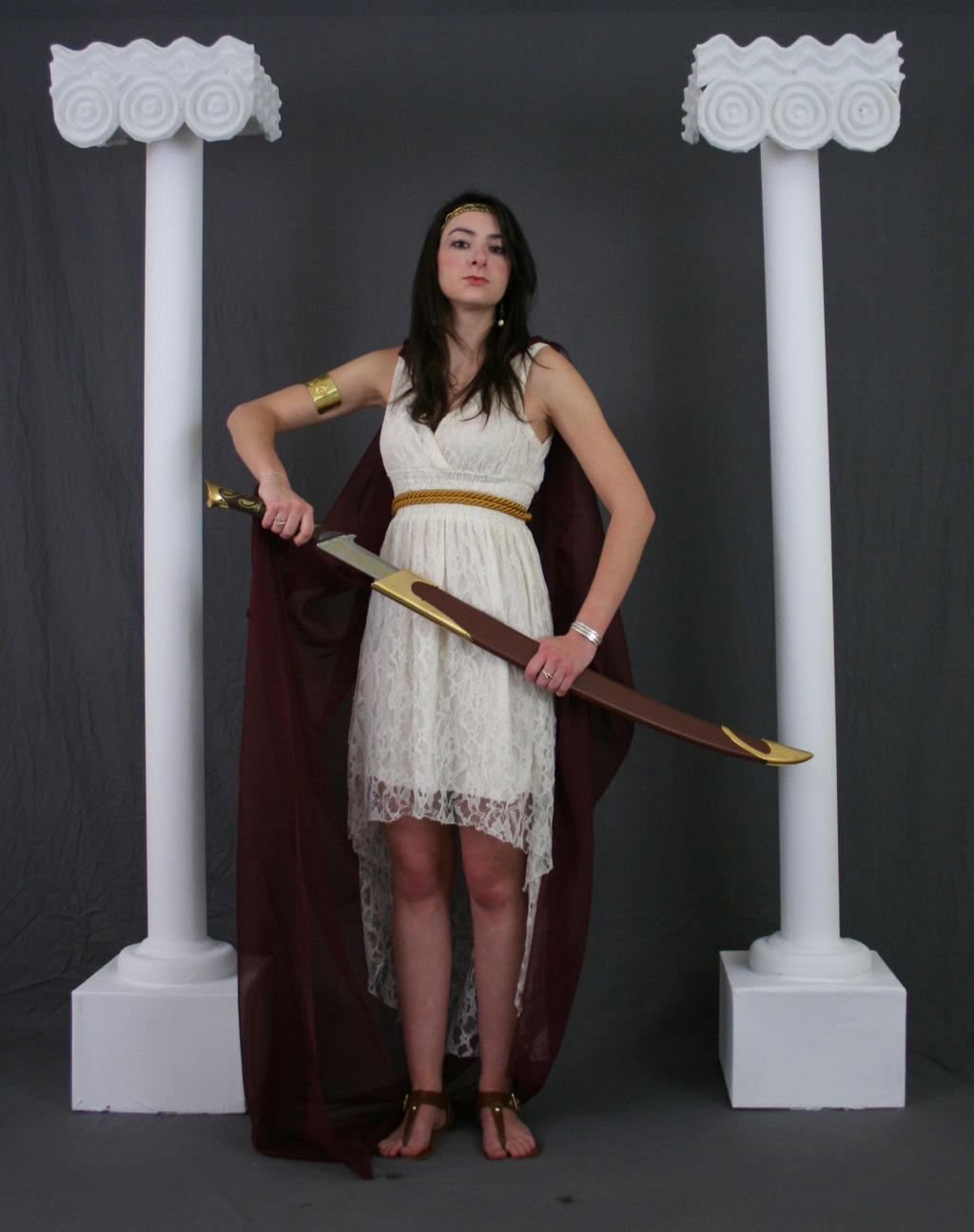 a full shot of a young woman dressed in an offwhite lace highlow dress and burgundy cape, holding a sword. she is standing between two white columns with decorative capitals, reminiscent of ancient greek or roman architecture. the background is a simple grey backdrop. the woman has dark hair and is wearing gold jewelry. her expression is serious and poised. the overall aesthetic is reminiscent of a mythological character or a historical reenactment.