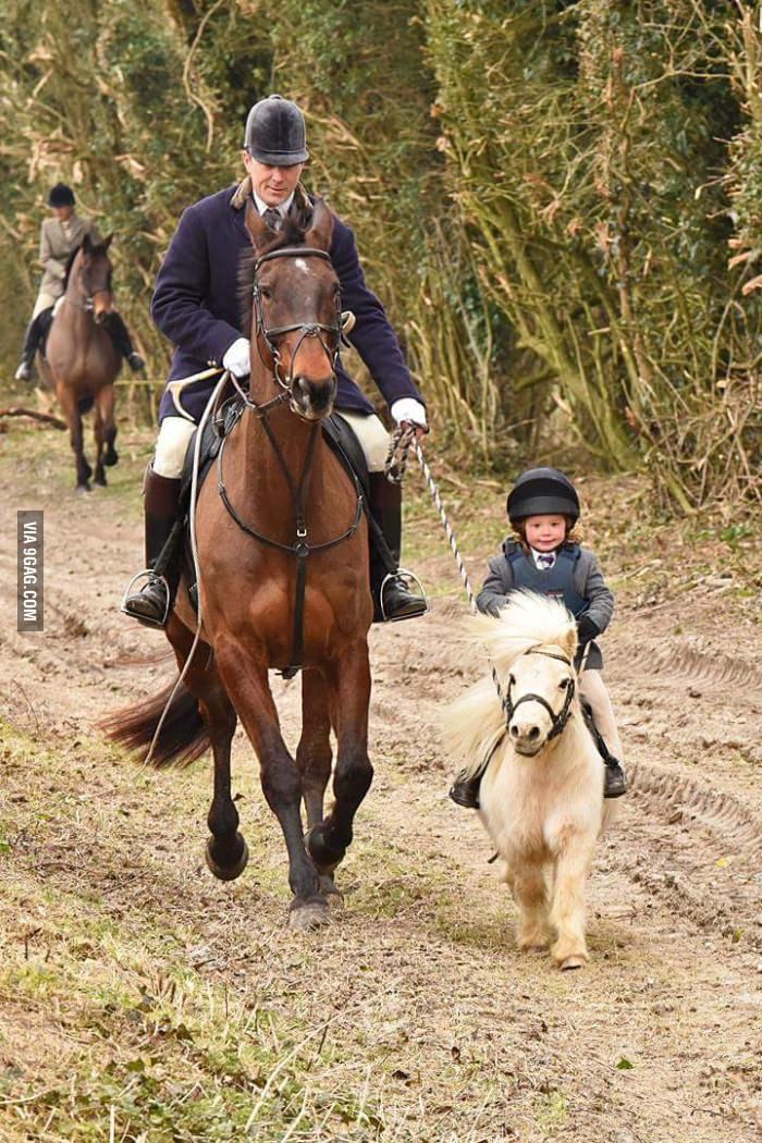 a man is riding a brown horse, and a young child is riding a small, lightcolored pony. the man is holding a rope that is attached to the pony's bridle. they are on a trail in the woods, with trees in the background. another rider on a horse is further back. the man is wearing a helmet, jacket, boots, and gloves, as is the child. both are wearing riding helmets. the horse and pony are both equipped with bridles and saddles.