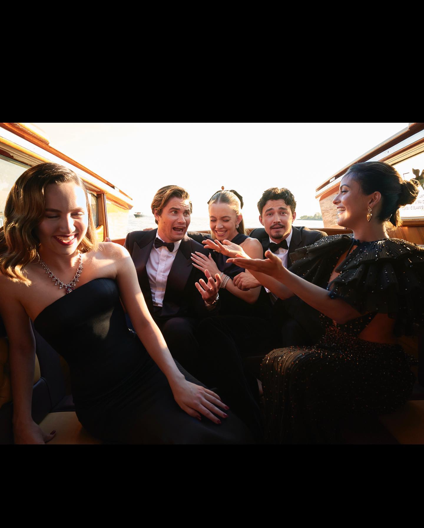 a group of elegantly dressed young men and women are sitting in a gondola in venice, italy.  the women are wearing long, dark evening gowns, while the men are in tuxedos. they appear to be happy and laughing, enjoying themselves during what seems to be a film festival or similar highprofile event. the sunlight creates a warm, golden hour ambiance.  the scene is luxurious and celebratory.