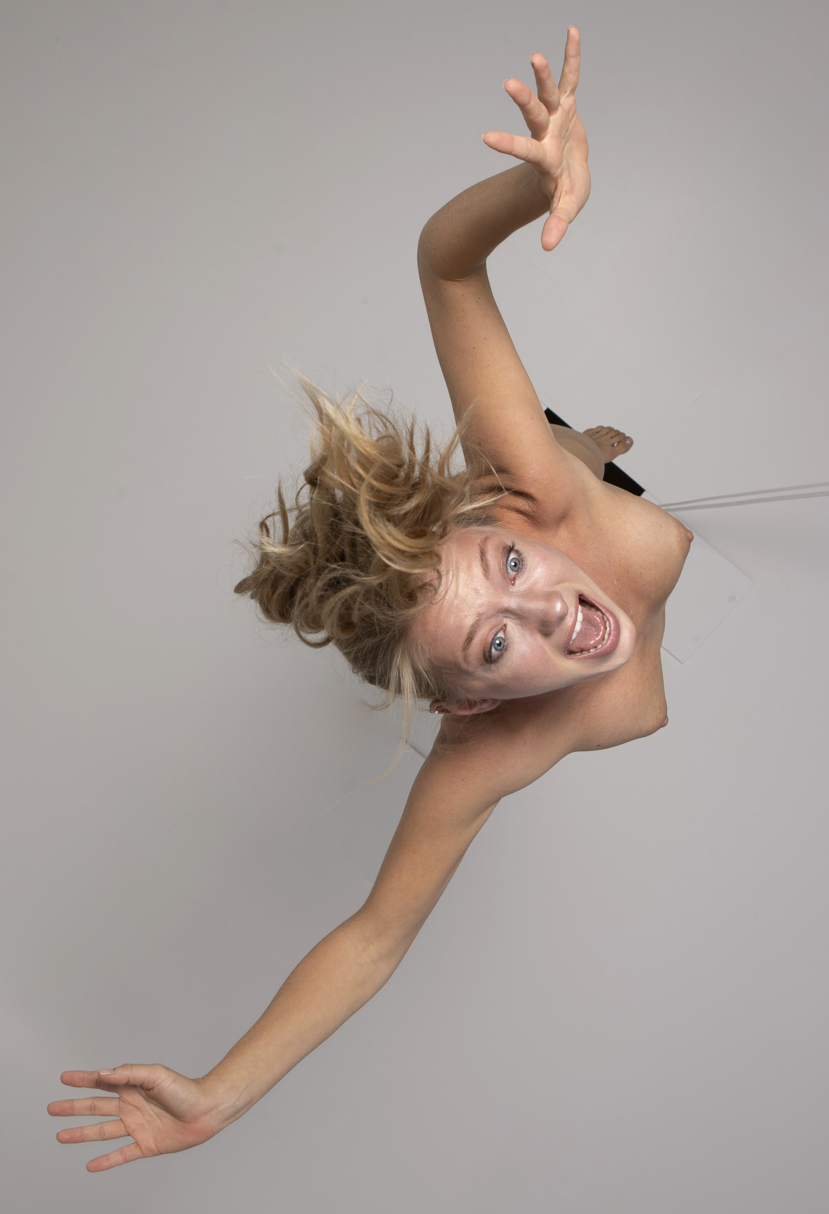 a woman with blonde hair is hanging upside down, performing some form of aerial acrobatics or dance. she has her arms extended and is looking up with her mouth open in an expressive pose. she appears to be in a studio with a white background, possibly suspended by a thin wire. she is wearing a dark top.