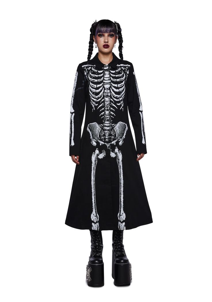 a woman with long black hair styled in pigtails is wearing a long black dress with a white skeleton printed on it. she's standing in a studio with a white background and is looking at the camera. she's wearing black platform boots with silver studs on the soles. she's dressed in a punk or goth style and could be going to a halloween party or an event.