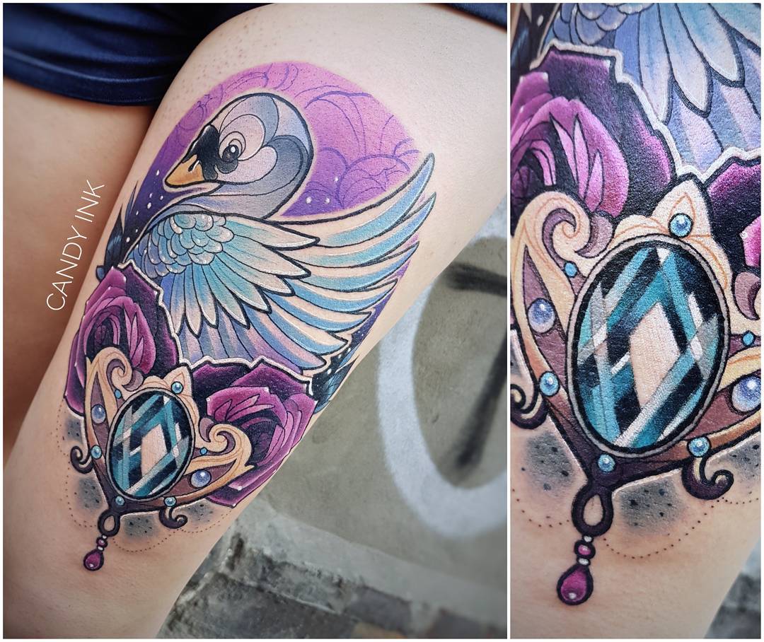 a tattoo of a swan with blue and purple wings on a right thigh. the swan is above a jewelencrusted  broach with a blue and white gem. the broach is partially obscured by purple roses with green leaves.  the background of the tattoo is purple with white dots.