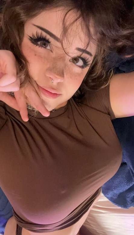 a selfie of a woman wearing a brown shirt. she has brown hair and is wearing a necklace and nose ring. she is looking at the camera.