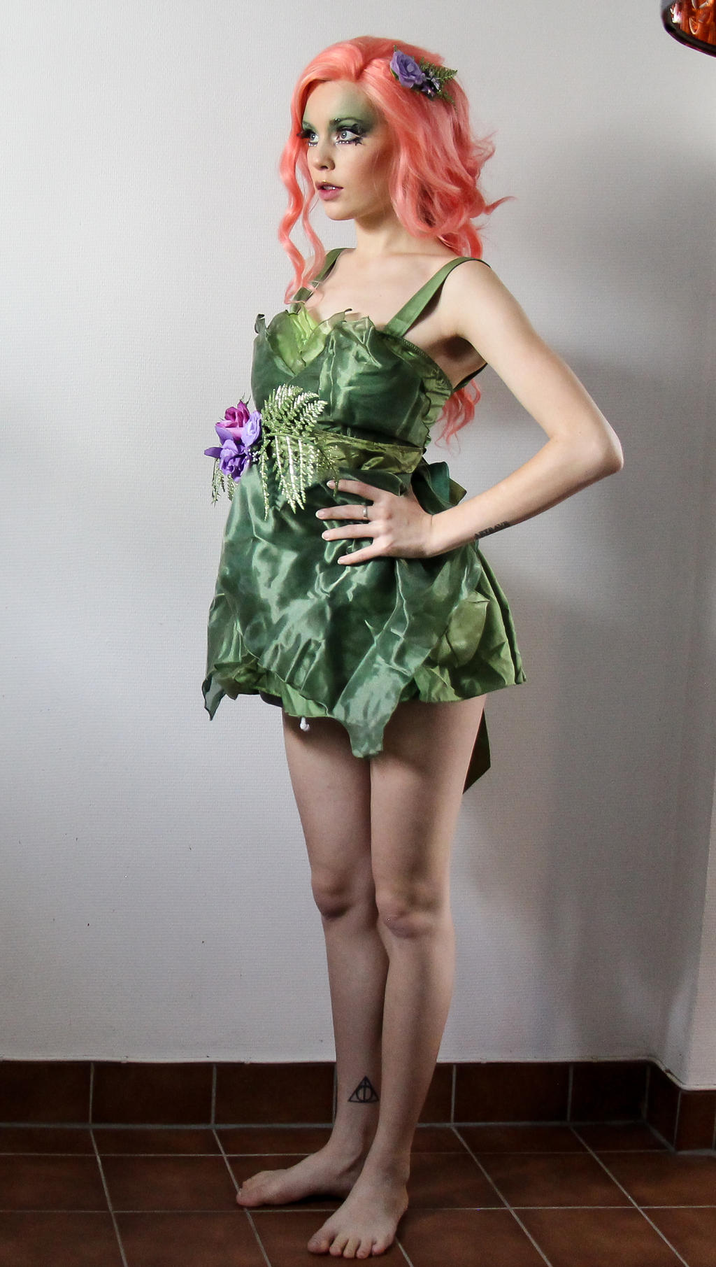 a fullbody shot of a young woman with bright pink hair styled in loose curls, wearing a green, floraladorned dress reminiscent of poison ivy. she is posing against a white wall with a brown tile floor visible. she has a subtle makeup look and is displaying a confident posture.  a small deathly hallows symbol tattoo is visible on her left ankle. the overall aesthetic is whimsical and elegant, suggestive of a fantasy or cosplay character.