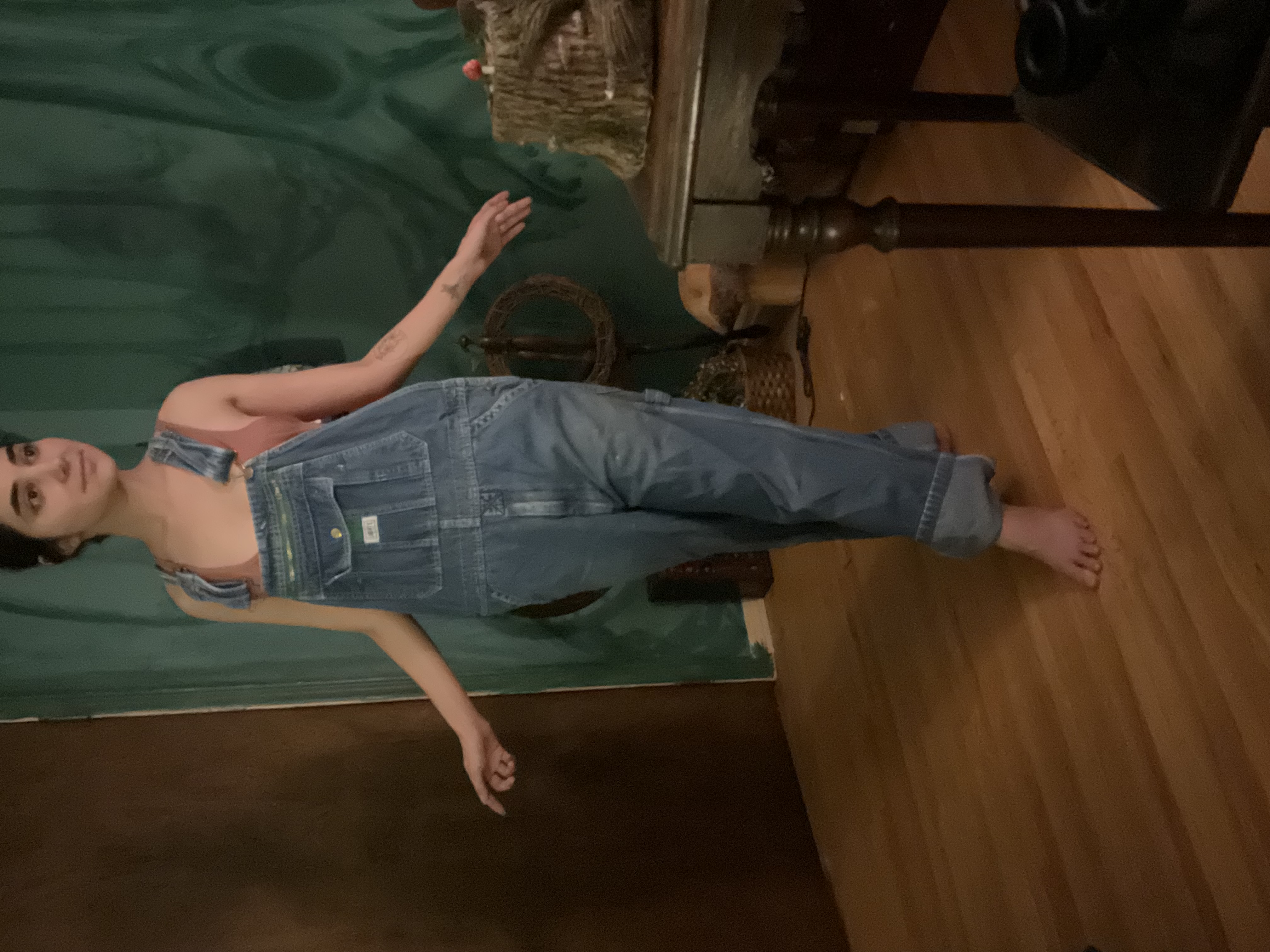 a fullbody shot of a woman with long brown hair wearing a pair of lightwash denim overalls. she's barefoot and standing on a hardwood floor in a room with a green tapestrycovered wall, a wooden side table visible behind her. the overall aesthetic is casual and relaxed.