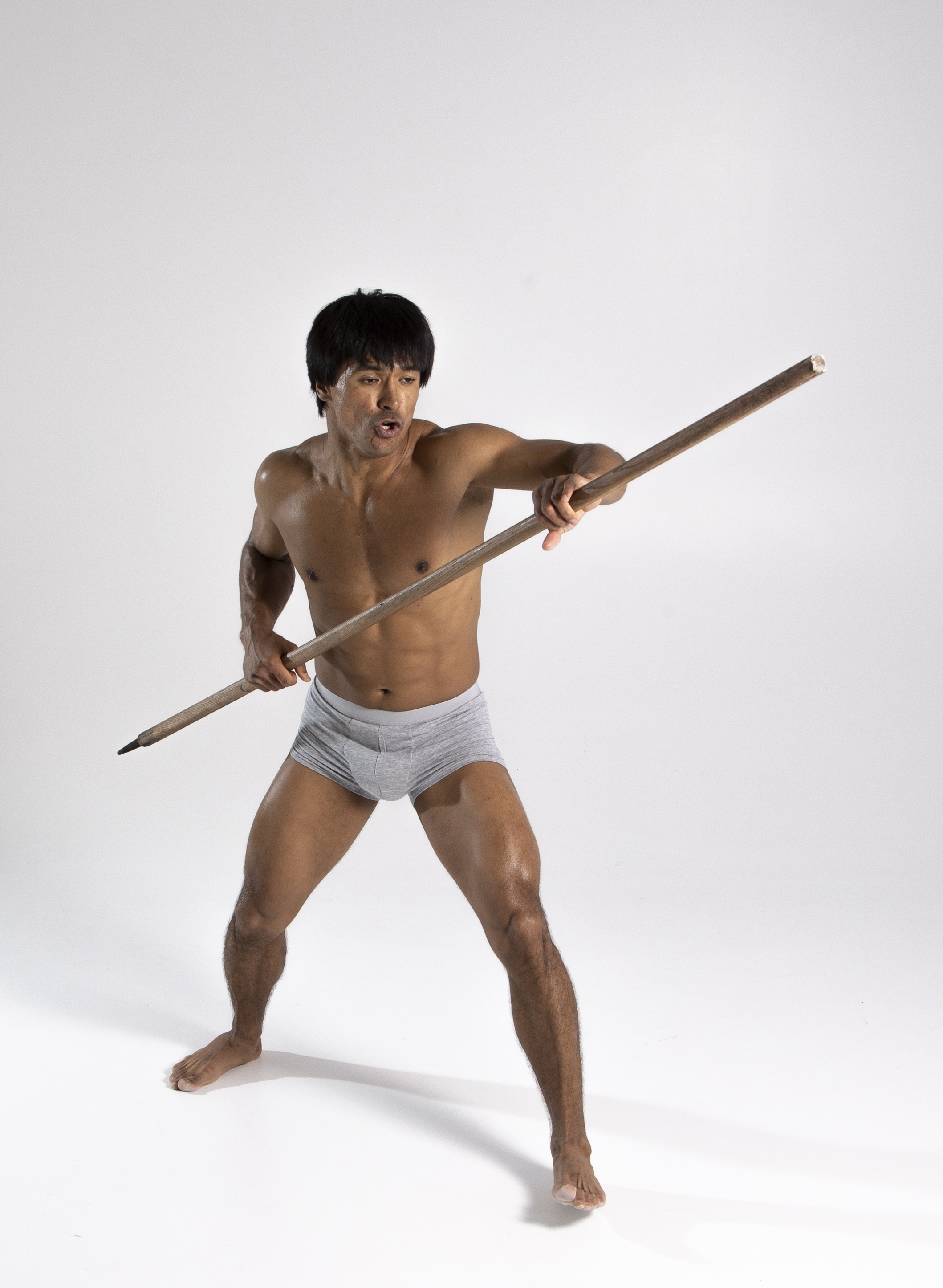a shirtless, muscular man is holding a long wooden stick resembling a spear in a defensive or attacking posture. he is barefoot and wearing grey underwear. the man is standing against a plain white background in a studio setting. his expression suggests focus and intensity.