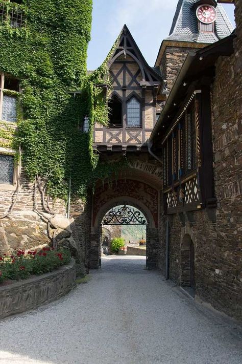 the image shows a stone archway, the entrance to a castle.  the building is made of stone and has ivy growing on it.  there are flowers on the left side of the archway.  the archway leads to a courtyard. the photo was taken on a sunny day.