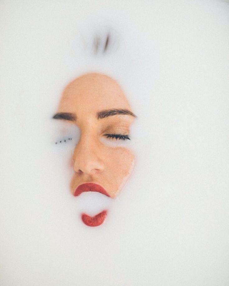 a woman's face is submerged in a milk bath, with only her eyes closed and red lips visible. the white milk creates a soft and ethereal backdrop, highlighting the woman's beauty and serenity. the image evokes a sense of relaxation, indulgence, and selfcare.