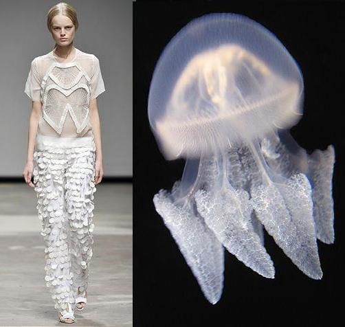 a fashion model wearing a white dress walks down a runway. the dress is made of delicate fabric and has a scalloped design. the model is standing on a black runway with a black background. to the right of the model is a jellyfish. the jellyfish is also white and transparent. the model and the jellyfish both have a delicate and ethereal quality.  the model is standing in the left side of the image. the model's white dress looks like an upsidedown jellyfish.