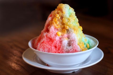 a bowl of shaved ice with red, green, and yellow layers is sitting on a white plate. the shaved ice has corn kernels mixed in.