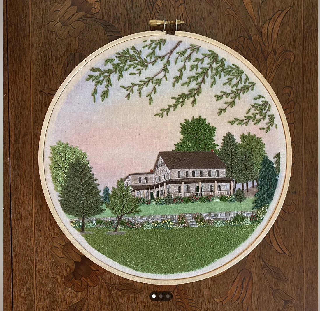 the image shows a piece of embroidery art in an embroidery hoop. the embroidery depicts a landscape featuring a twostory house with a front porch, surrounded by trees and a garden. the scene is set against a backdrop of a sunset sky. the artwork uses various shades of green, brown, and pink.