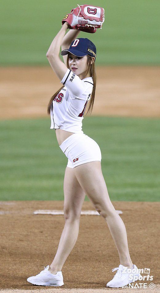 a young woman, dressed in a cropped white baseball uniform with red and blue details, stands on a baseball field preparing to throw a pitch. she's wearing a baseball glove on her right hand and has her left arm raised. the field is visible behind her, with green grass and brown dirt. the lighting is bright and sunny, indicating a daytime game.
