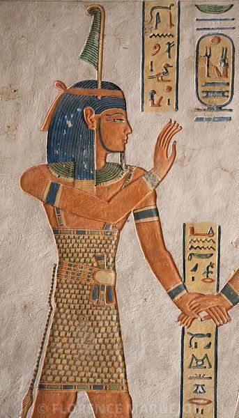 the image shows a painting of an egyptian pharaoh in profile view, facing right. he wears a traditional kilt and headdress with a feather. his skin is painted a reddish brown, his hair is blueblack, and his clothing is yellow and gold. he is gesturing with his hands toward columns of hieroglyphs. the background is a pale cream color. the painting is illuminated by bright, natural light.