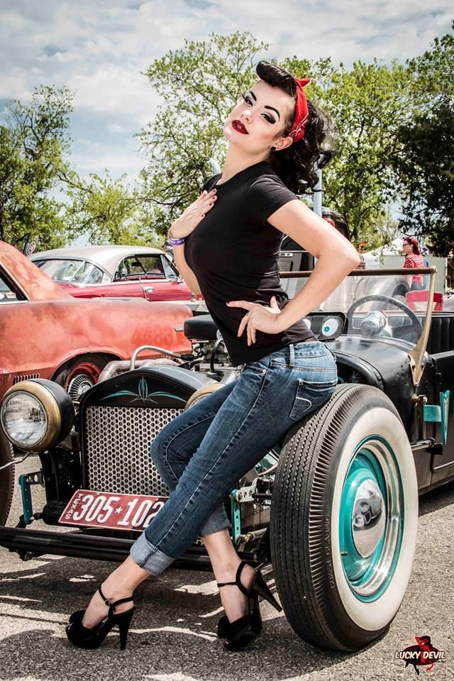 a pinup girl in a red bandana, black top, and jeans poses seductively on a vintage hot rod.  the car is black with teal accents and large whitewall tires. the setting appears to be an outdoor car show with other classic cars visible in the background under a partly cloudy sky. the woman's makeup is dramatic and her hair is styled in a classic 1950s fashion. the overall image evokes a retro, rockabilly aesthetic.