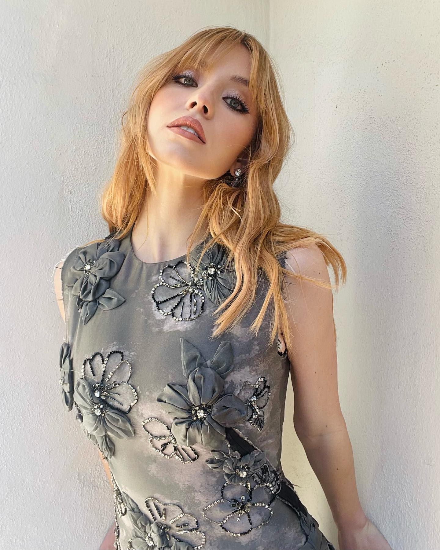 loseup portrait of a young woman with long blonde hair and bangs, wearing a stylish sleeveless grey dress adorned with dark grey fabric flowers and crystal embellishments. the dress is fitted and the design is intricate and detailed. the woman is posing confidently and her expression is alluring. she has subtle makeup and is wearing small earrings. the backdrop is a simple white wall.