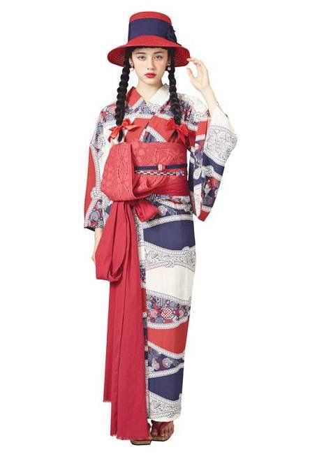 a fulllength studio shot of a young woman wearing a colorful kimono and a matching hat. the kimono is predominantly red, navy blue, and white, featuring a floral pattern throughout. a red sash is tied around her waist, and she is also wearing a widebrimmed hat in red and navy blue. her hair is styled in two braids, and she is posed against a plain white background.