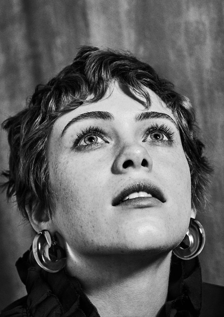 a black and white closeup portrait of a young woman with short, curly hair. she is looking upwards, and her face is framed by large hoop earrings. the image is high contrast, with dramatic lighting emphasizing the texture of her skin and hair.  she has freckles and a serene expression. the overall style is elegant and sophisticated.