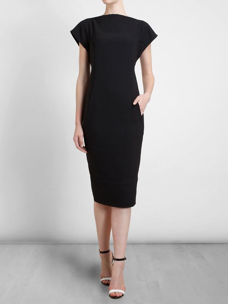 a woman is standing in front of a white background wearing a black midi sheath dress and sandals. she has one hand in the side pocket of the dress.