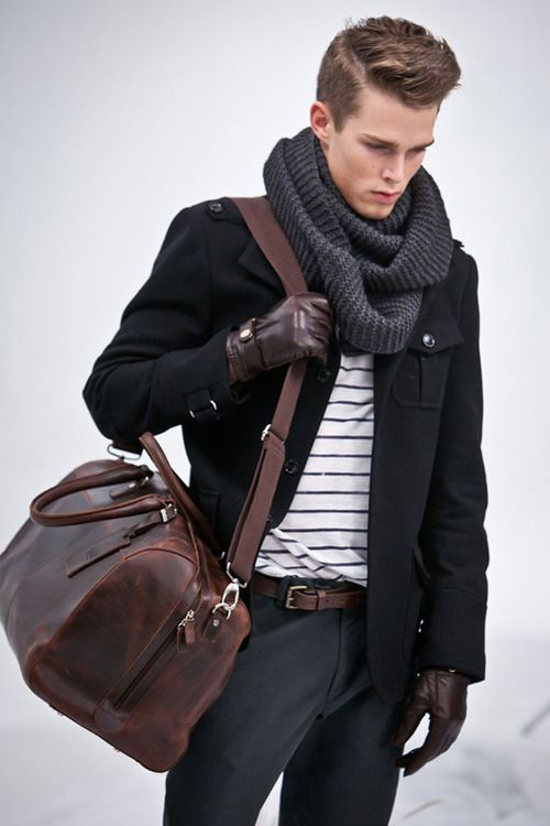 a man in a black coat, grey scarf, and brown leather gloves is carrying a brown leather bag. he's standing in an outdoor setting, and the background is a light, snowy white. he's wearing a white striped shirt under his coat, giving a casual, stylish vibe. the leather bag hangs from his shoulder and is accented by silver hardware. the man has a slight smile, making him appear confident and approachable. the overall tone of the image is stylish and confident, suggesting a man who knows his fashion sense.