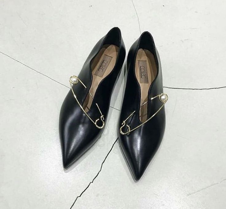 a closeup shot of a pair of black leather pointedtoe flats.  each shoe features a gold safety pin with a small pearl detail, elegantly placed across the vamp. the shoes are positioned on a light grey floor, and the brand label is visible inside. the image focuses on the unique detail of the safety pin accent, highlighting the shoes' stylish and minimalist design.