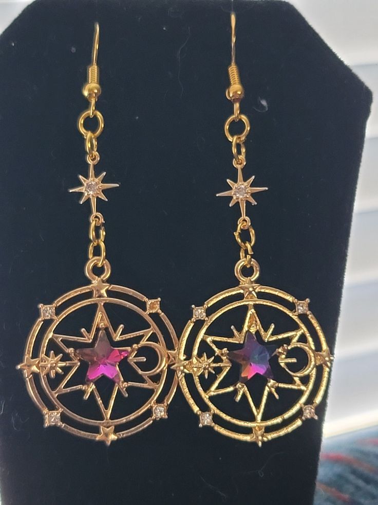 a closeup shot of a pair of gold dangle earrings.  the earrings feature a circular design with a central, large, iridescent purple star surrounded by smaller stars and crescent moons.  the circular design is detailed and intricately crafted. the earrings hang from gold chains which feature a smaller star charm halfway down. the earrings are displayed against a black background.