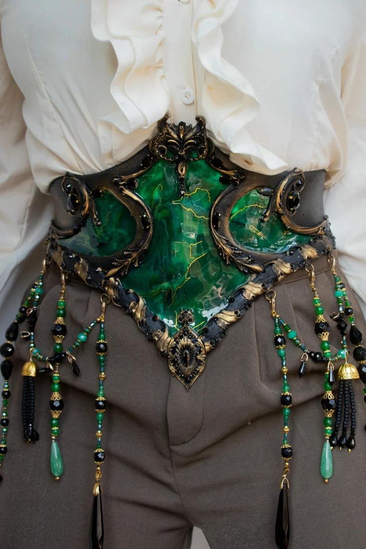 this is a closeup photo of a woman's waist adorned with a luxurious emerald green and gold belt. the belt is intricately designed with ornate details, featuring a swirl pattern and gold accents, and it sits over a brown skirt or trousers. the belt is accented with dangling black and emerald green beads that add a touch of bohemian charm to the look. this is a perfect example of a bold and eyecatching accessory that could complete a steampunk, fantasy, gothic, victorian, or cosplay look.