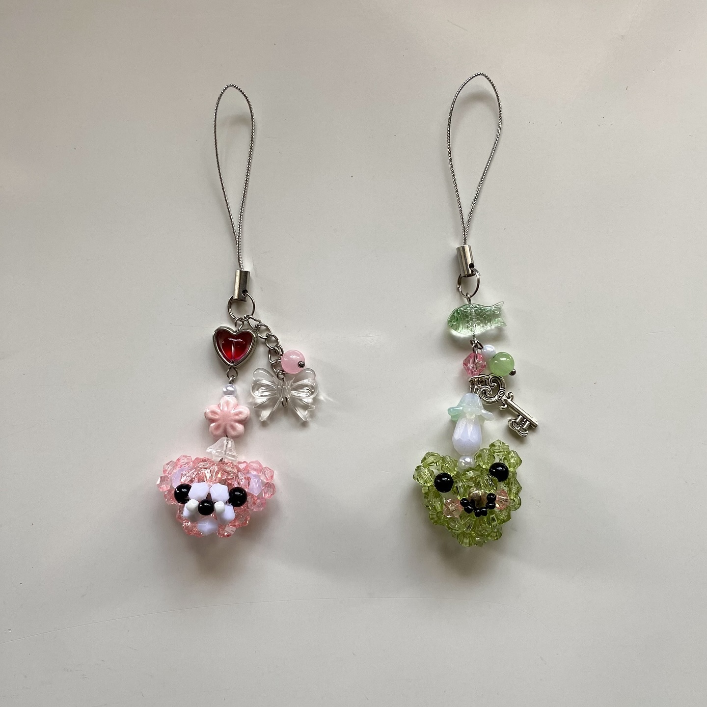 the image shows two handmade beaded phone charms. one charm features a pink beaded dog face with black and white accents, a pink flower, a red heart, and a clear bow. the other charm features a green beaded frog face, a key charm, a mushroom charm, and various green and pink beads. both charms are attached to silver phone straps.