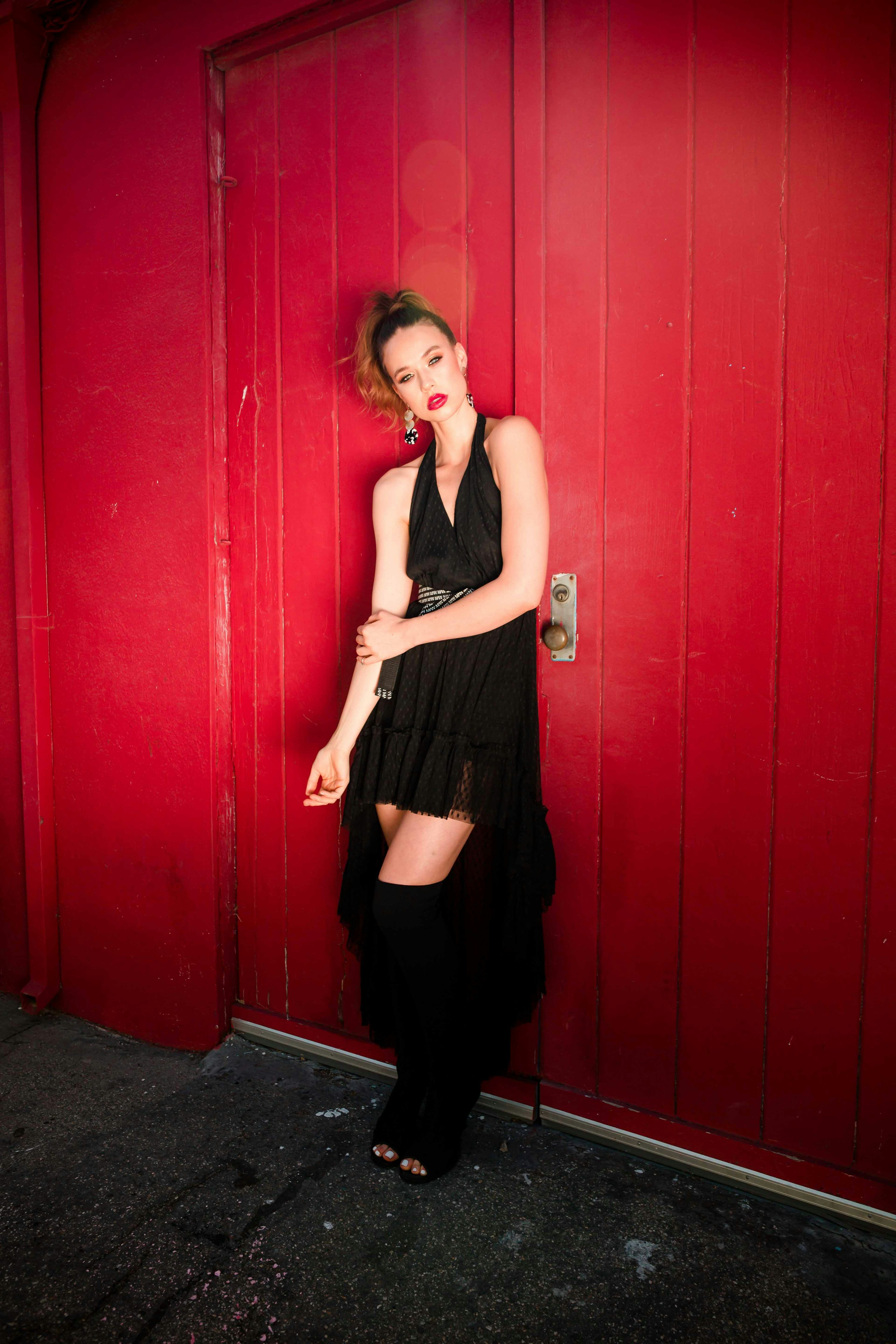 a fullbody shot of a fashion model standing against a vibrant red door. she's wearing a black, highlow dress with lace details, paired with black thighhigh boots. her makeup is striking, featuring bold red lipstick, and she's accessorized with delicate earrings. the model's pose exudes confidence and style, creating a captivating image against the backdrop of the red door. the lighting is dramatic, highlighting her features and the textures of her clothing and the door.