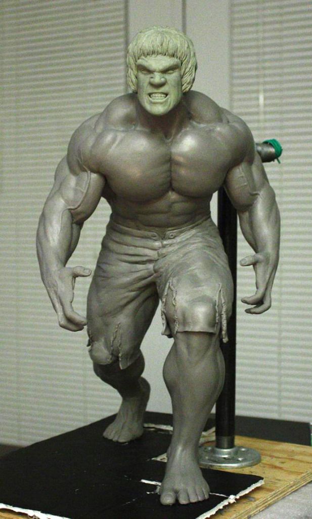 a gray clay sculpture of the hulk, a popular marvel comics superhero, stands on a wooden platform in an art studio. the hulk is depicted in a dynamic pose with his fists clenched and teeth bared in anger. the sculpture captures the character's immense muscular physique and iconic green skin, although it is currently unpainted. the studio setting is evident from the plain background and artificial lighting.