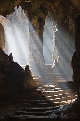 a photo shows a dark cave with stone steps leading up and away. sunlight streams into the cave, creating light rays and strong shadows. the cave appears to have stalactites or other cave formations hanging from the ceiling. the stairs look ancient and manmade, giving the impression of an underground passage.