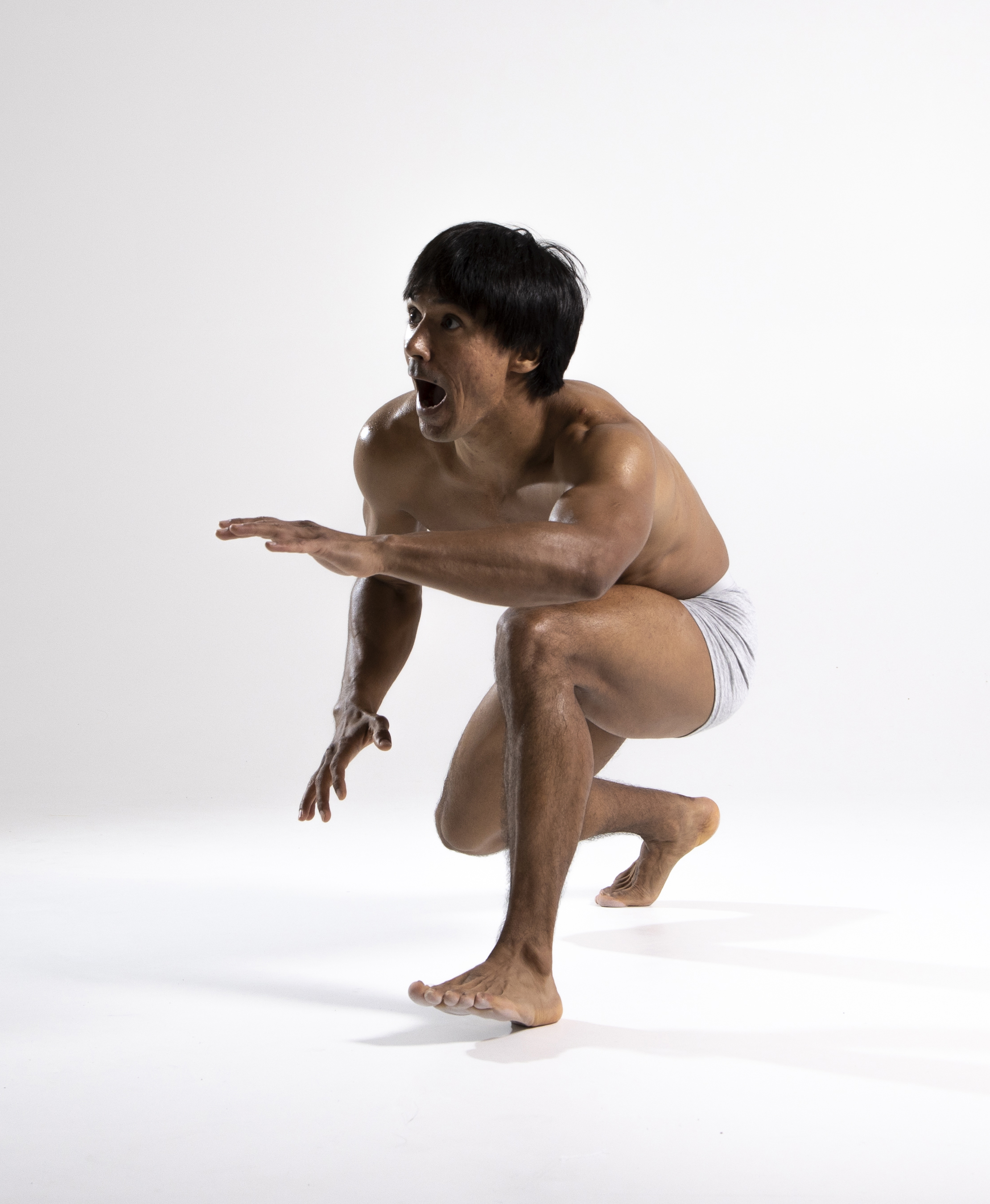 a muscular, barechested man with dark hair is crouching on a white background. he is wearing gray shorts and is barefoot. his mouth is open in a scream, and his expression is one of intense emotion. the photo is a studio shot with even lighting.