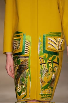 a woman is wearing a vibrant yellow coat adorned with intricate green and brown embroidery. the embroidery features a floral motif, creating a striking visual contrast against the bright yellow fabric. the model's confident pose and the coat's elegant design highlight the beauty and sophistication of the garment.