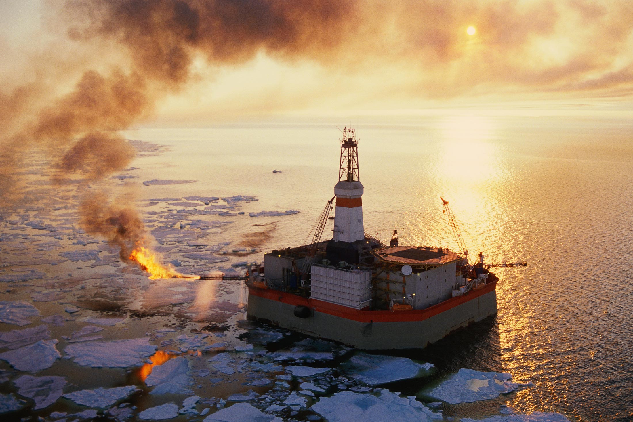 an oil rig sits in the middle of an ocean covered in ice floes. smoke and flame billow from the rig. the sun sets in the background, casting a warm glow on the scene.