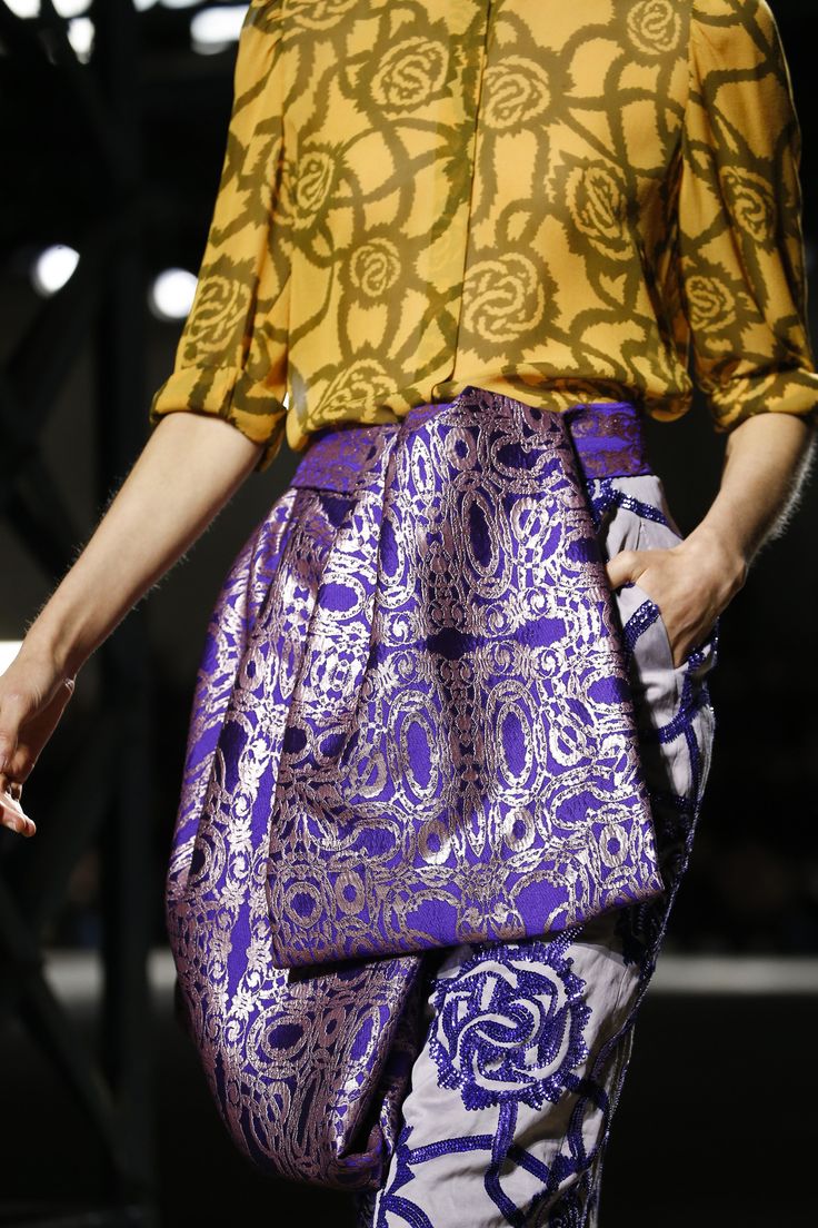loseup view of a woman's fashion model showcasing a stylish outfit. the ensemble features a mustard yellow silk blouse with a distinct floral pattern. the blouse is paired with a purple brocade skirt or pants with an intricate woven design in metallic gold thread. the skirt's design is a draped style with a wraparound effect. the model is partially visible, emphasizing the luxurious texture and detail of the clothing. the backdrop is dark and blurred, focusing attention on the attire. the overall impression is one of sophisticated and unique design within women's high fashion.