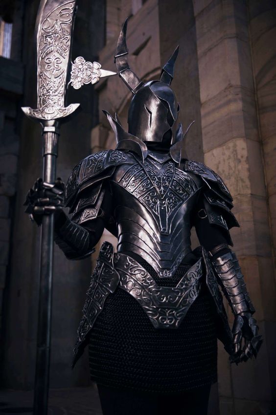 a person in elaborate, dark, ornate cosplay armor stands poised in front of a gothic stone archway. they hold a large, metal halberd and their helmet features large horns. the overall aesthetic is reminiscent of the black knights from the dark souls video game series.  the lighting is soft and natural, casting shadows that accentuate the details of the armor.