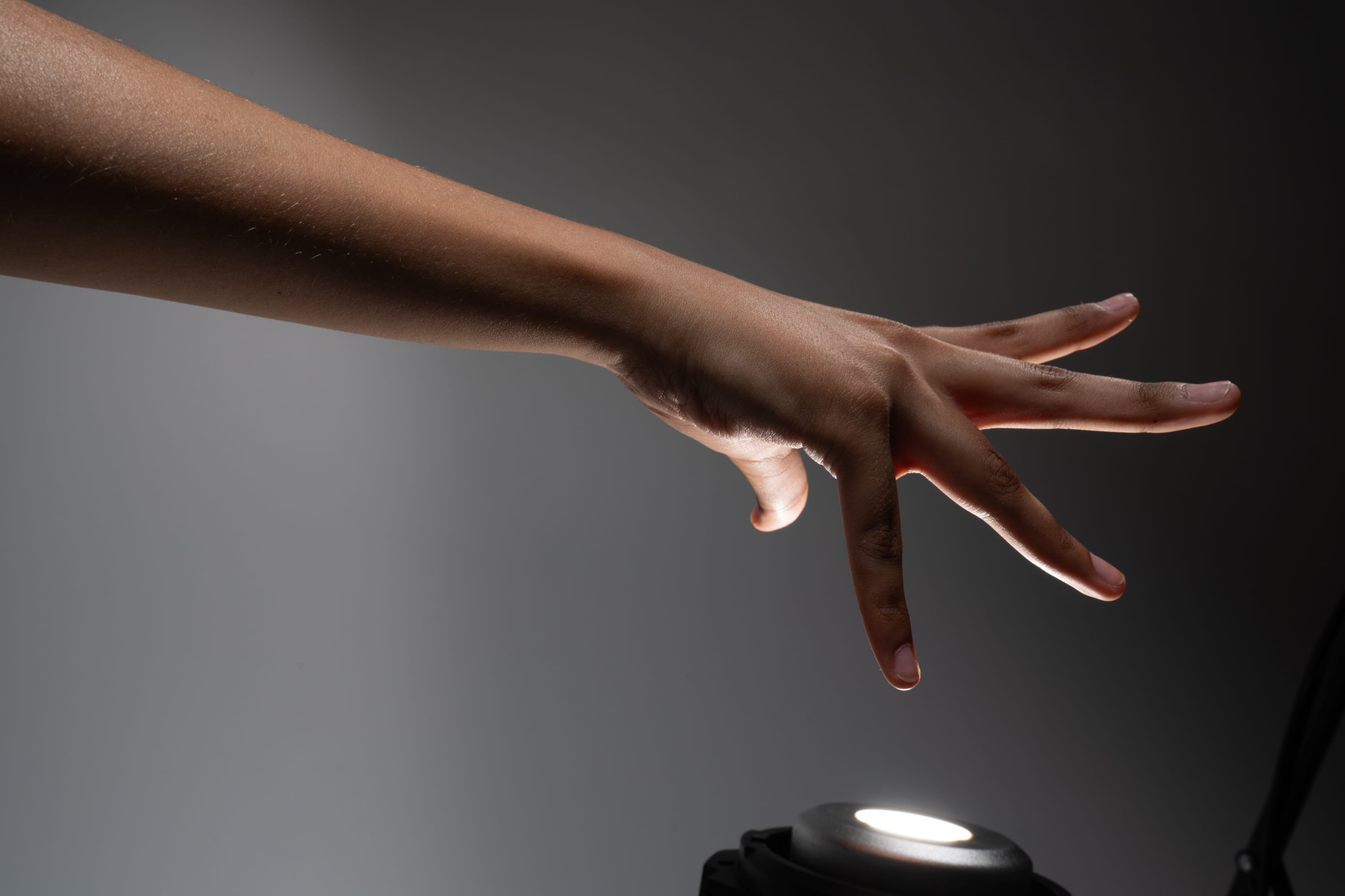 a hand reaches toward a small, circular light source. the light casts a dramatic shadow across the hand and arm, highlighting the contours and textures of the skin. the background is a dark gray, creating a stark contrast with the illuminated hand. the overall composition evokes a sense of mystery and intrigue.