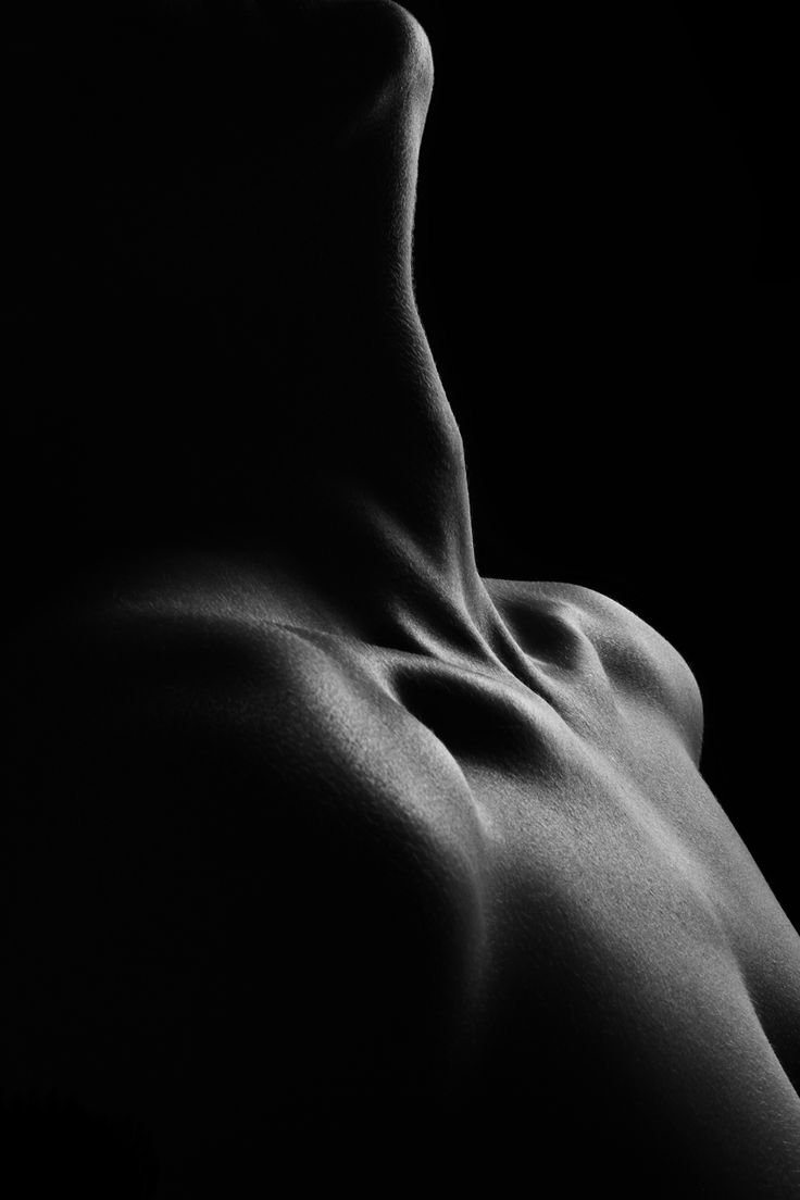 the image is a black and white closeup photograph of a woman's nude torso. the lighting is dramatic, using strong contrasts of light and shadow to highlight the curves and texture of the skin. the focus is on the graceful lines of the neck, shoulders, and upper chest, emphasizing the form and anatomy of the body.  the overall effect is sensual and artistic, with an abstract quality due to the minimal background and use of shadow. the photo is reminiscent of fine art nude photography.