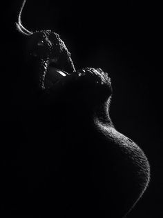 the image is a dramatic black and white closeup shot of a pregnant woman's silhouette.  the lighting is strongly backlit, creating a high contrast image where most of the woman's form is in shadow, highlighting the curves of her pregnant belly and back. the texture of her skin is subtly visible where the light catches it. the overall mood is sensual and artistic, with a focus on the form and shape of the body.