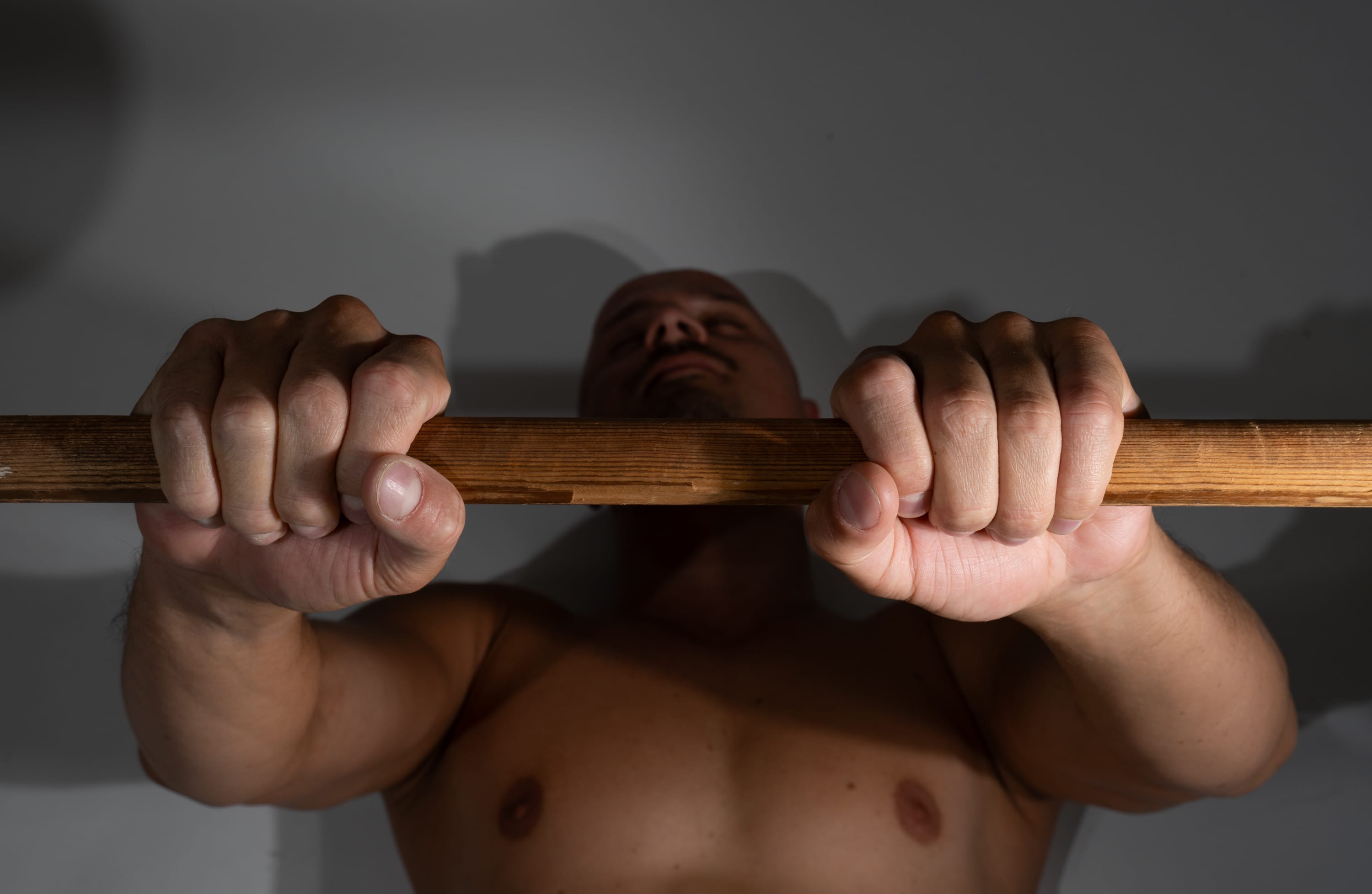 a shirtless muscular man is seen from a low angle perspective as he grips a wooden bar, likely in the process of performing a pullup exercise. his face is turned upwards, focusing on his effort, and the lighting emphasizes the muscular definition of his arms and upper body. the background is a simple, dimly lit grey wall, keeping the focus on the man's physical exertion.