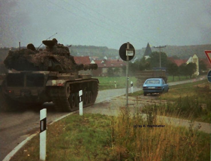 a photograph depicting a camouflaged tank driving down a wet road in a rural german village. the tank is covered in mud and appears to be moving slowly. a small blue car is visible in the background. the scene is overcast, and the overall mood is somber. the photo appears to be a vintage print, possibly from the 1980s, as suggested by its style and quality. the photo is credited to r. hochstatter.