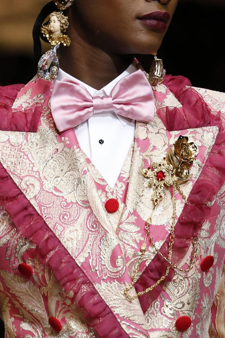 loseup shot of a model wearing a pink and gold brocade suit. the suit features a doublebreasted jacket with ruffled trim, a pink bow tie, and ornate gold jewelry including large earrings and a brooch with a rose motif.  the focus is on the intricate details of the clothing and accessories, showcasing the high quality and luxurious nature of the design.  the image suggests a fashion show or editorial photoshoot.