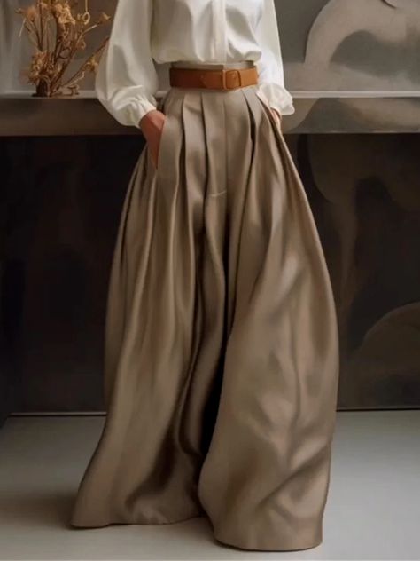 a woman is standing, wearing wide leg, highwaisted pleated trousers, a white shirt and a brown belt. her hands are in the pockets of her trousers. the background is neutral and minimalist. the lighting is soft and the image is taken at eye level.