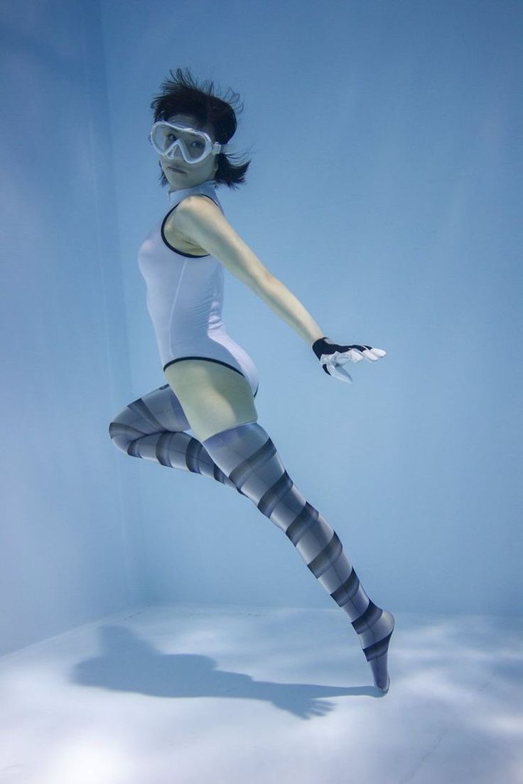 a woman in a white onepiece swimsuit and patterned tights poses underwater in a pool. she wears white swimming goggles and black fingerless gloves. her pose suggests movement and athleticism. the background is a uniform, bright blue, and the lighting is artificial, highlighting the woman and creating a sense of depth despite the monochrome backdrop.